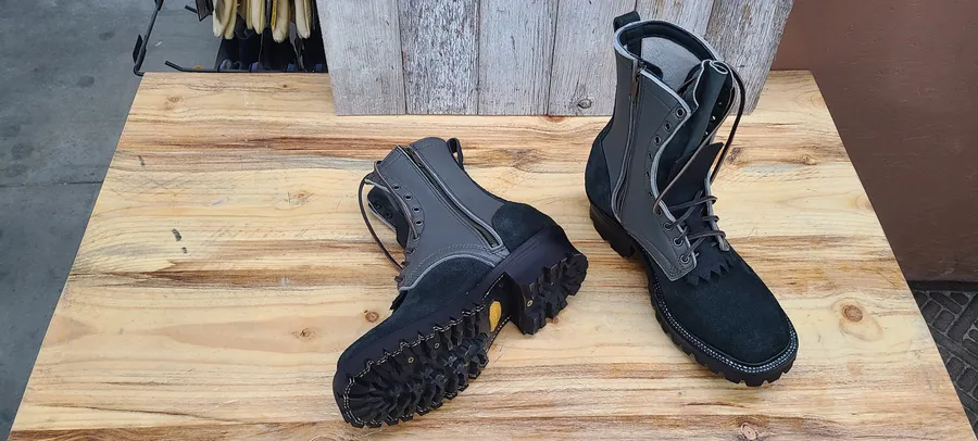 A pair of tall black fire boots on a pine bench in front of weathered barn boards: rough-out feet, smooth grey-lined shafts, fringed kilties and lugged soles, one boot lying on its side to show the tread.