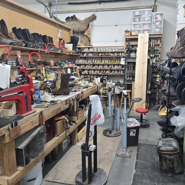 A workshop bench crowded with tools and part-built lace-up boots, a red Champion stitcher at the near end, shelves of boot lasts across the back wall and finishing wheels on the right.