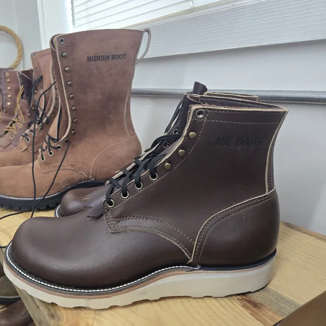 Finished boots on a wooden shelf: a tall rough-out lace-up stamped HIDDEN BOOT beside a brown smooth-leather boot with a white wedge sole stamped ABE BOOTS.
