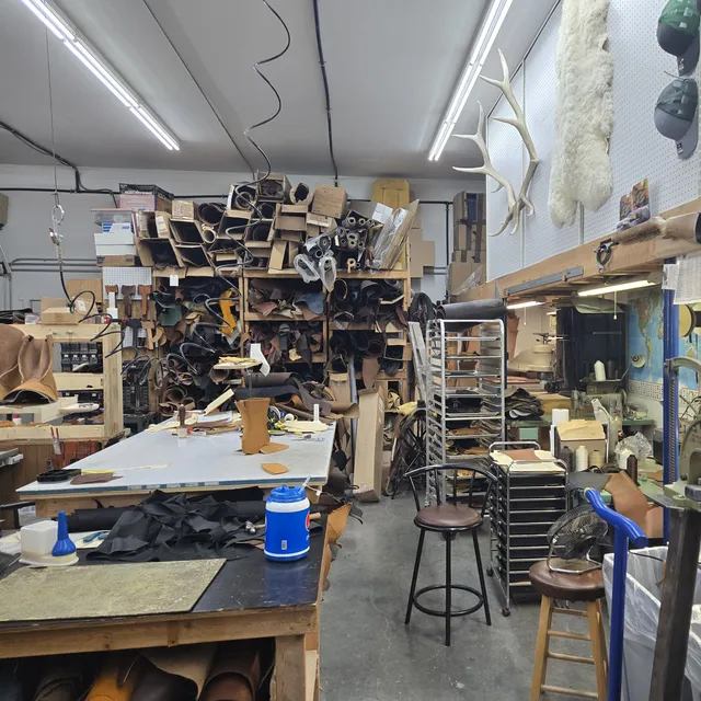 A workroom with a cutting table in front of floor-to-ceiling racks of rolled leather hides, a rolling tray rack, and sewing machines along the right wall.