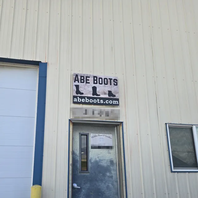 The door of a tan steel building with an ABE BOOTS abeboots.com sign above it and an older Hiddin Boot & Leather card on the door. The street number and the address on the card are pixelated.