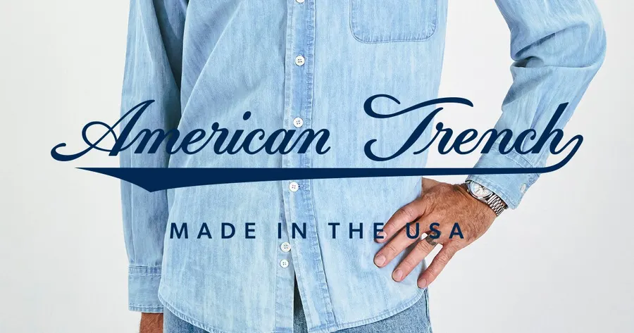 The torso of a person wearing a light blue button-down shirt with one hand on their hip, overlaid with American Trench lettering and the words Made in the U.S.A.