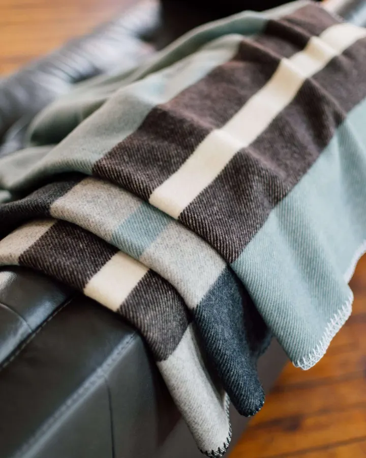 A sage-green, charcoal, and cream striped wool throw blanket folded over the arm of a dark leather chair on a hardwood floor.