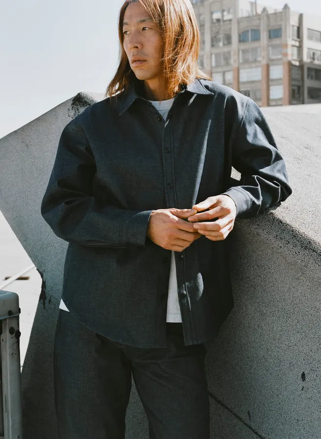 A man with long auburn hair leans against a concrete wall on a rooftop, wearing a dark grey denim overshirt buttoned over a white t-shirt.