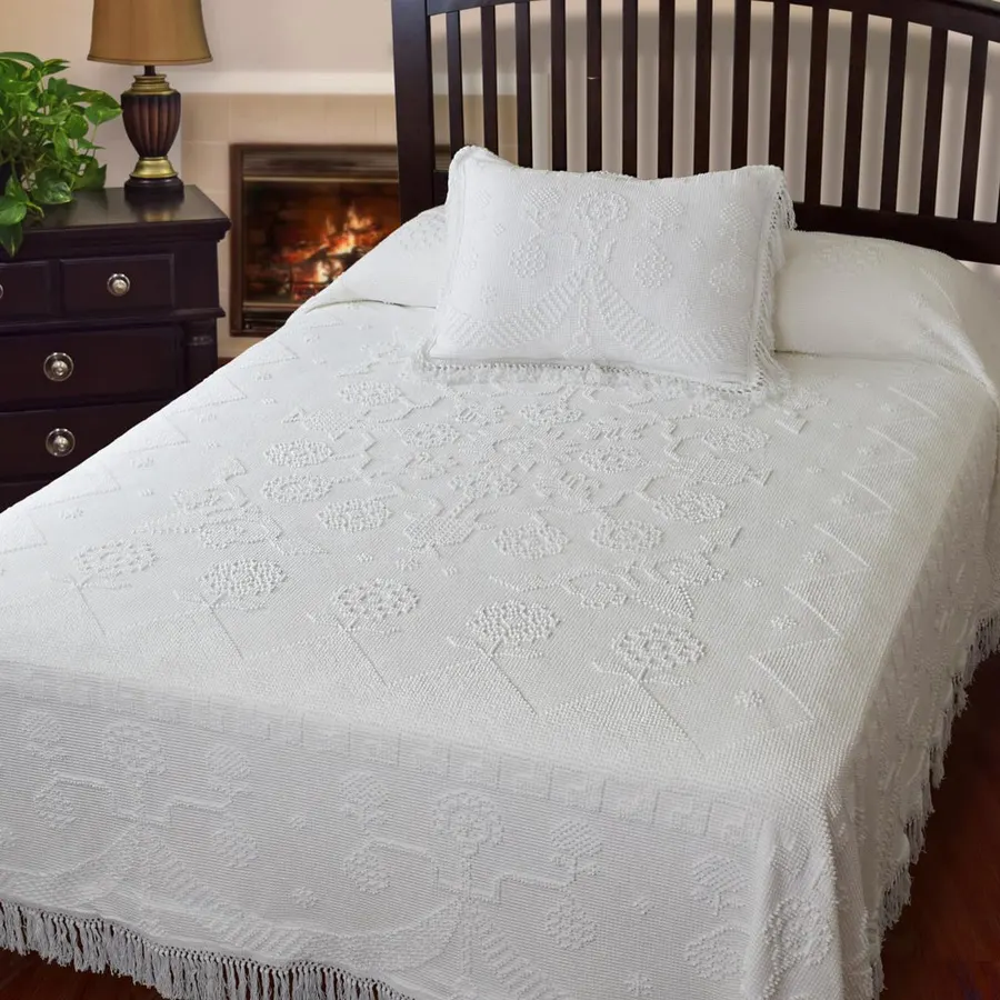 A white woven jacquard bedspread with a floral fringe-edge pattern, dressed on a bed with a matching pillow sham, dark wood headboard and nightstand behind.