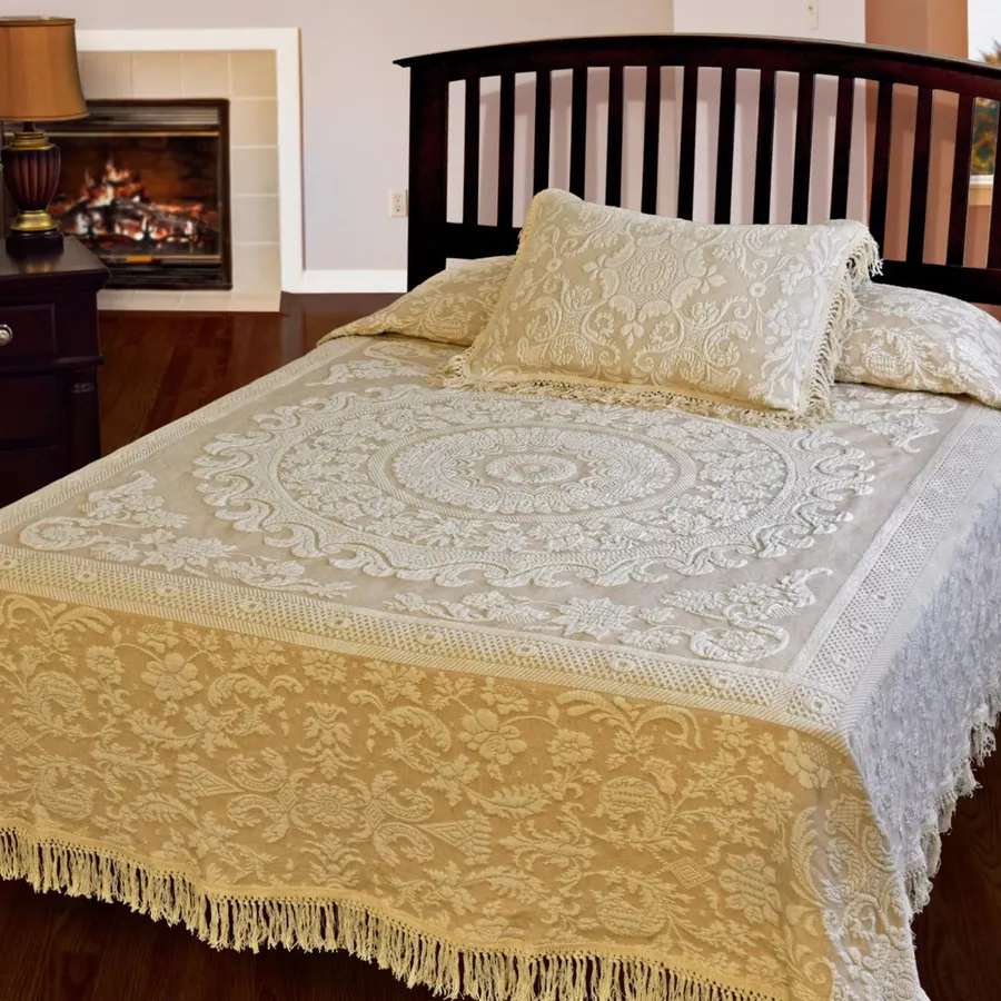 A cream matelasse bedspread with an ornate medallion and scroll pattern and fringe trim, dressed on a bed with a matching pillow sham, dark wood headboard and nightstand behind.