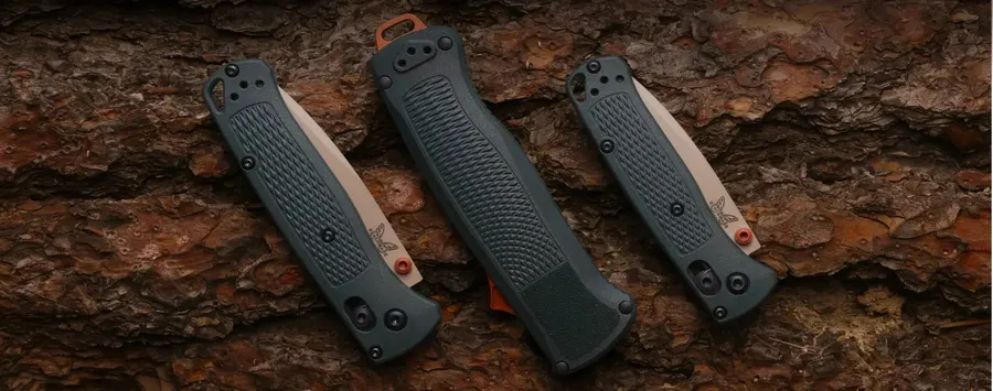Three closed Benchmade folding knives with dark gray handles and orange accents arranged diagonally against a dark, textured rock background.