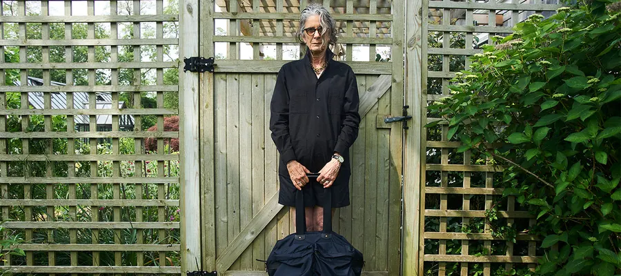 A gray-haired model in a dark coat and sunglasses stands in front of a wooden garden gate, holding a large navy bag.