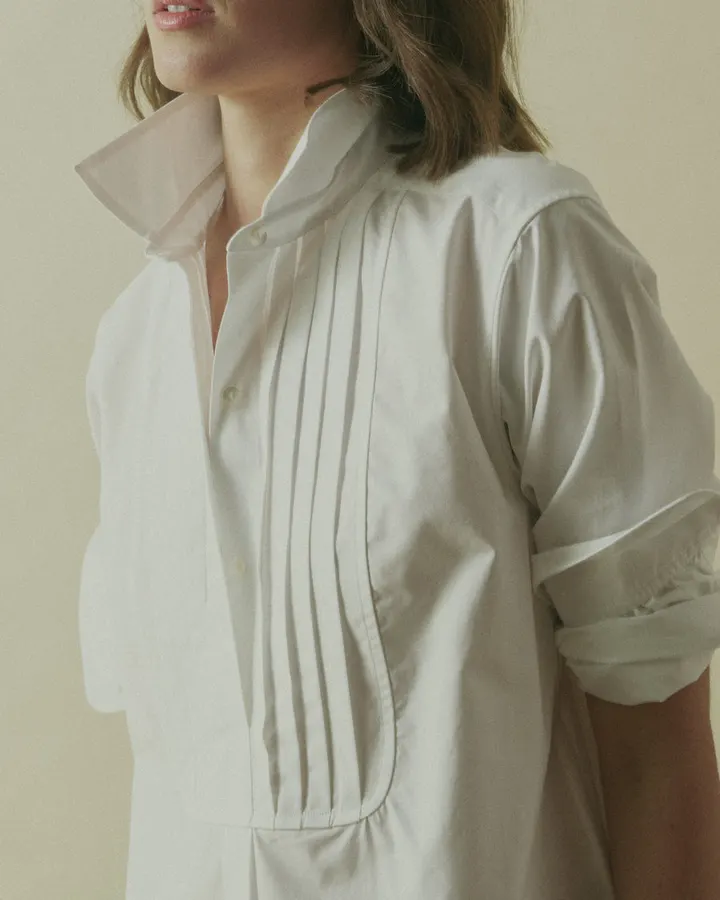 Close-up of a white poplin shirt's pintucked front bib and pointed collar on a model, sleeve cuff rolled at the wrist.
