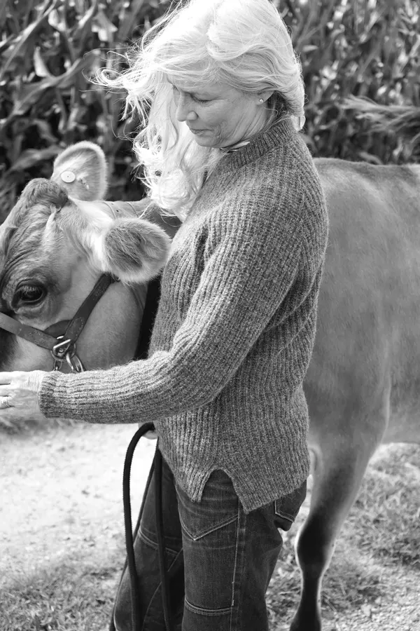 A black-and-white photo of a woman with long white hair wearing a chunky ribbed knit sweater, standing beside a cow in a cornfield.