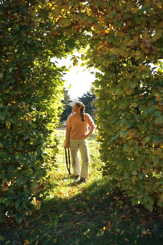 A woman in a peach knit sweater and cream trousers stands in a sunlit garden archway framed by autumn hedge foliage, holding a garden tool.