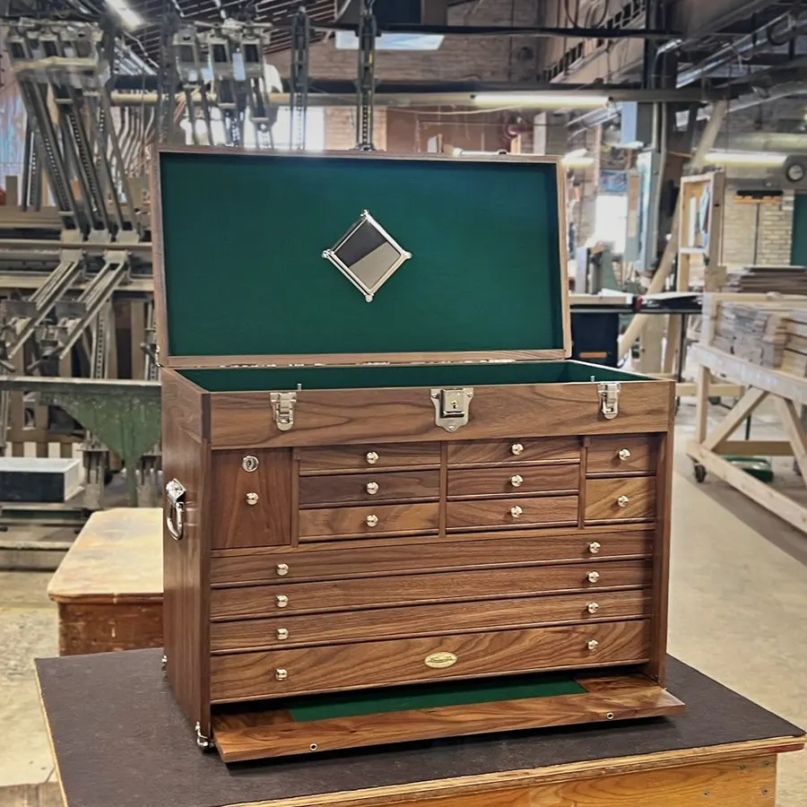 An open walnut tool chest with green felt-lined drawers and a mirror in the lid, standing on a table on a factory shop floor surrounded by woodworking machinery and lumber racks.