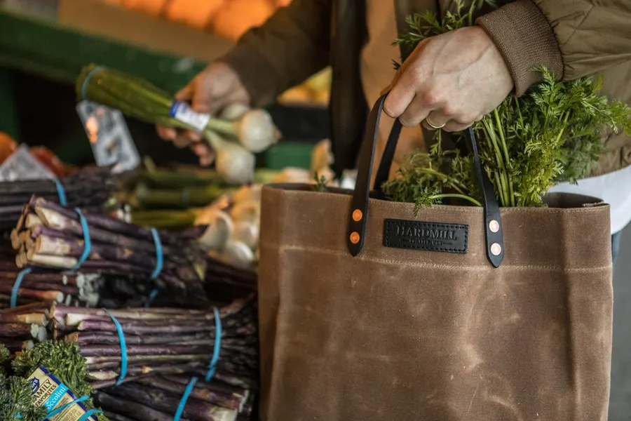 A hand holds open a brown waxed-canvas tote bag with black leather handles and a Hardmill patch, filled with fresh vegetables at an outdoor farmers market stall.