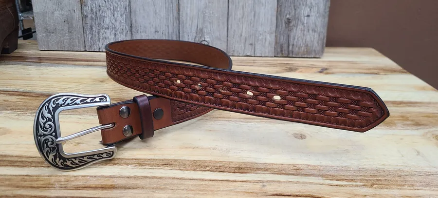 A brown leather belt stamped with a basket-weave pattern, coiled on a pine bench in front of weathered barn boards, its engraved silver buckle resting in the foreground.