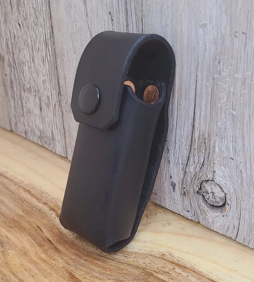 A black leather belt sheath with a snap flap, holding a multitool, leaning against weathered grey barn boards on a wooden bench.