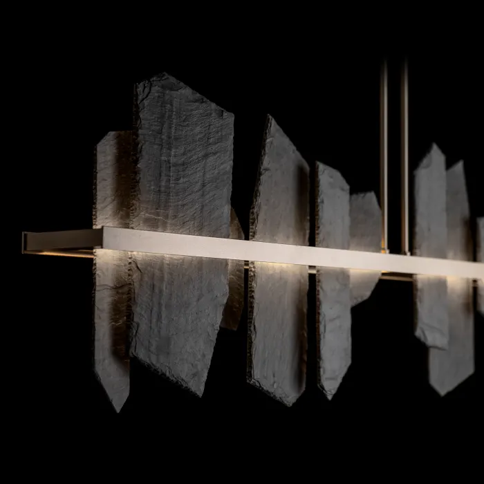 A linear LED pendant light with irregular slate-shard panels flanking a slim bronze bar, glowing warmly against a black background.