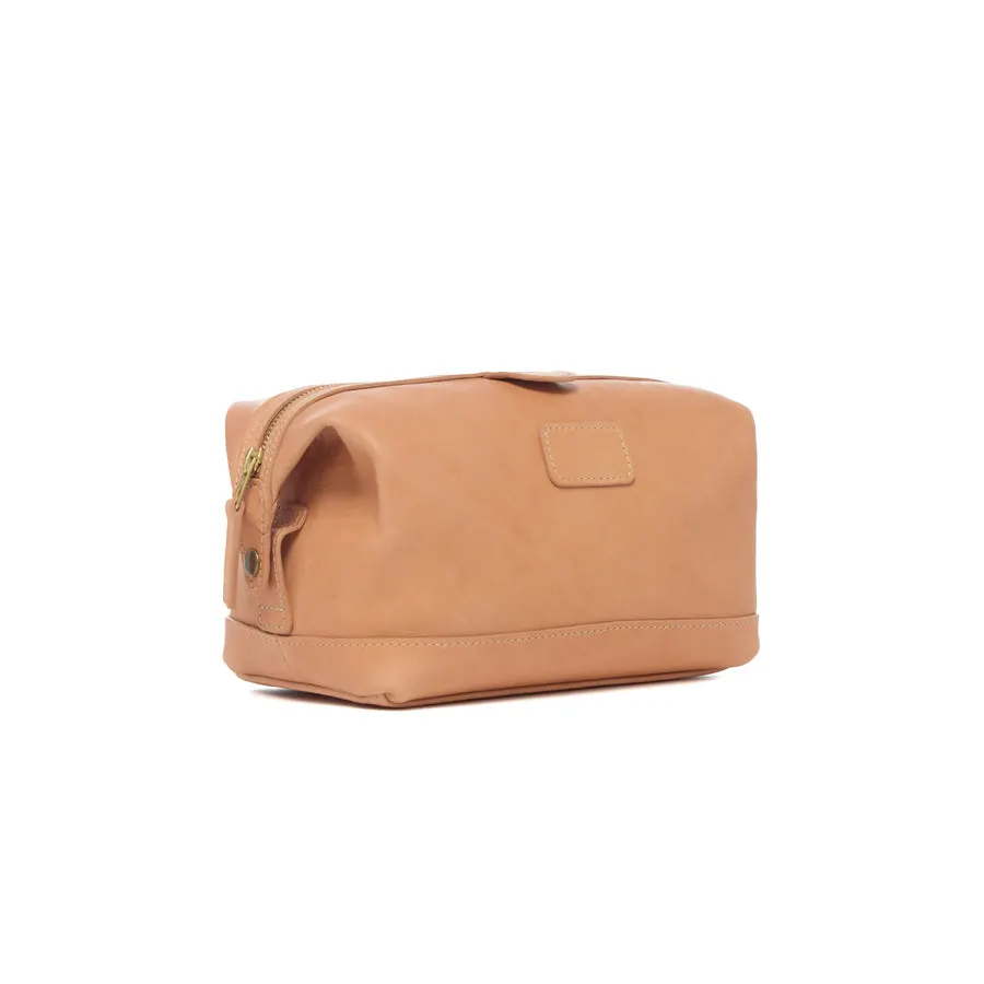 A tan leather dopp kit with a brass zipper and a blank leather luggage tag, shown at a three-quarter angle against a white background.