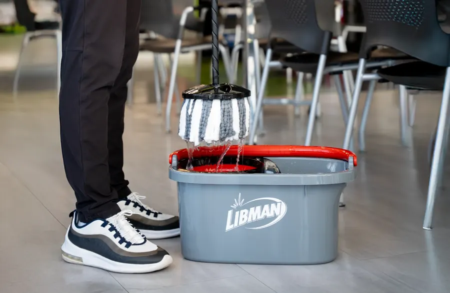 A gray Libman mop bucket with a red rim and white string mop beside a person wearing dark trousers and sneakers on a tiled floor.