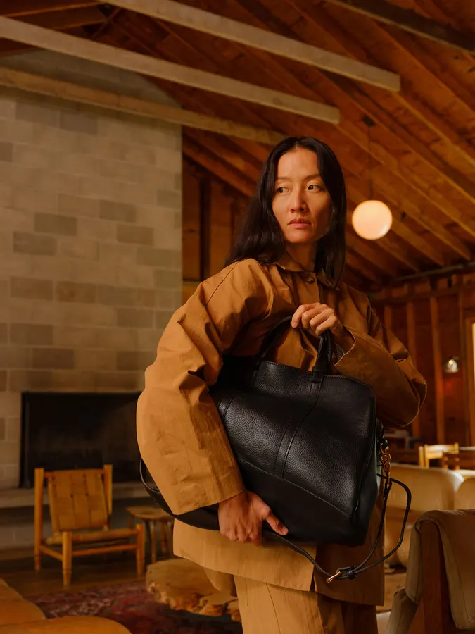 A woman in a tan jacket and trousers holding a black leather duffel bag against her chest, standing in a warmly lit wood-beamed room with a stone fireplace.