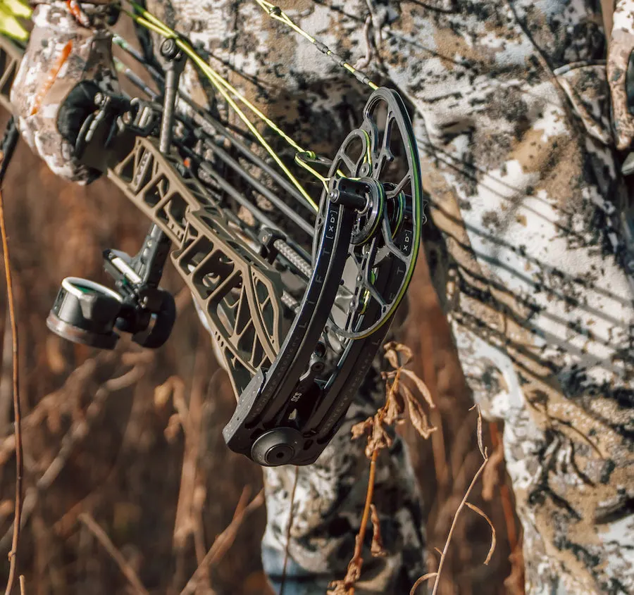 Close-up of a black compound bow held by a hunter in winter camouflage, aimed upward through dry brush.