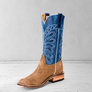 A single square-toed cowboy boot standing on a pale concrete floor against a white wall: a tan rough-out leather foot, a stacked leather heel, and a tall royal-blue shaft stitched with a white scroll pattern.