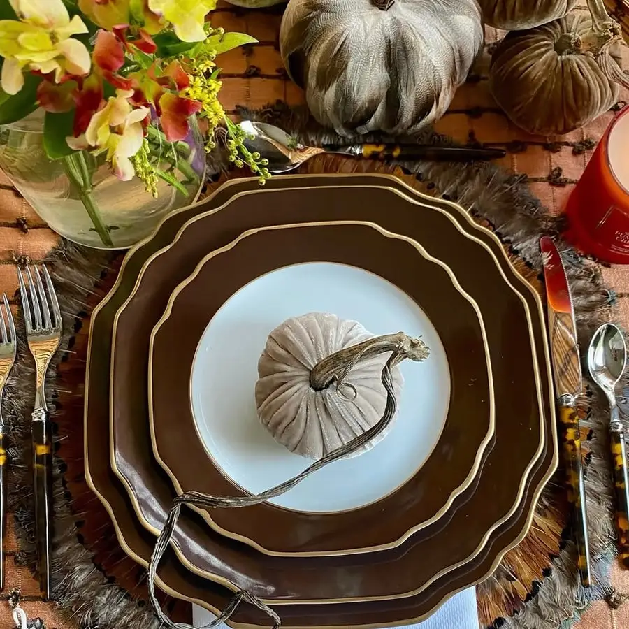 An overhead view of a formal fall table setting: a scalloped brown-and-gold Pickard charger and dinner plate topped with a white plate holding a small velvet decorative pumpkin, flanked by flatware, surrounded by fresh flowers and velvet pumpkins.