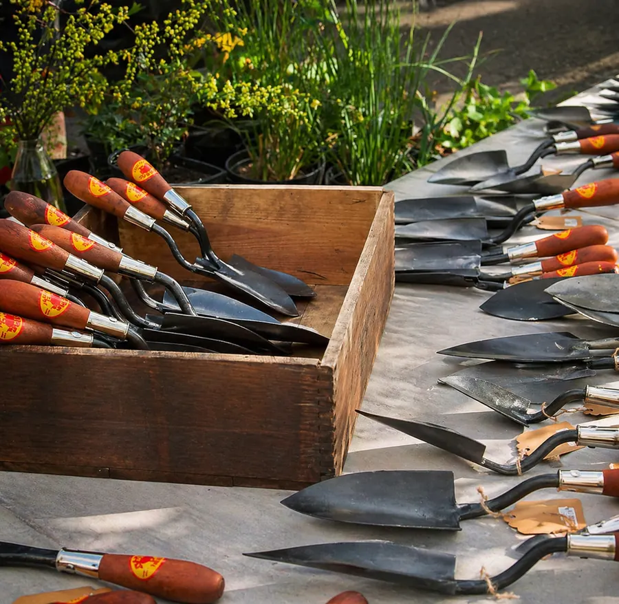 Hand-forged garden tools with wooden handles and Red Pig labels fill a wooden crate and lie on a table among green plants.