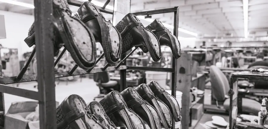 A black-and-white photograph of a boot factory floor: a metal rack in the foreground holds rows of unfinished cowboy boots upside down on their lasts, soles facing out, with workbenches, chairs and machinery stretching back under long fluorescent lights.