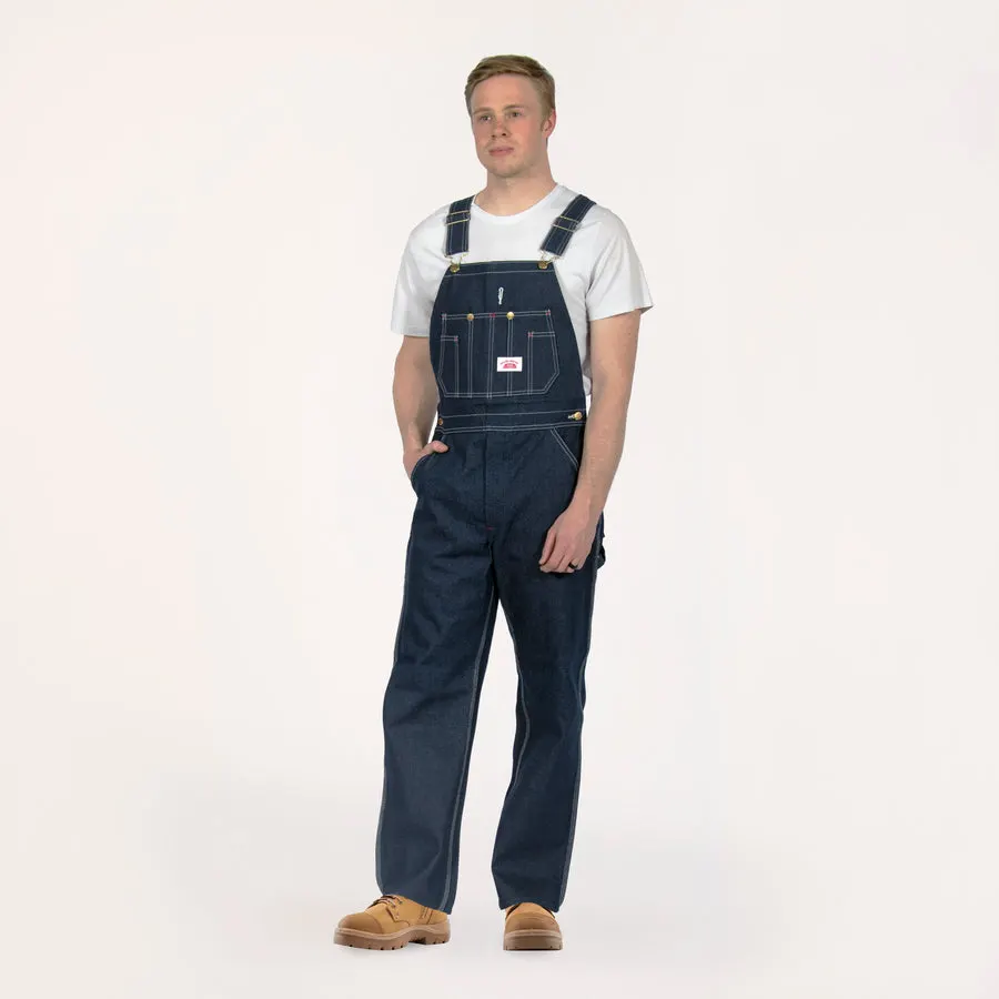 A young man wearing dark blue denim bib overalls over a white t-shirt and tan work boots, standing against a plain studio background.