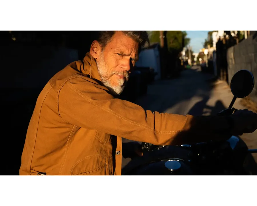 A bearded man in a rust-brown canvas work jacket glances back over his shoulder while gripping the handlebar of a parked motorcycle in an alley lit by low evening sun.