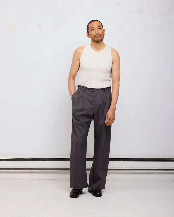 A man in a cream tank top and dark grey double-pleated wide-leg trousers, standing against a plain white wall with exposed pipe.