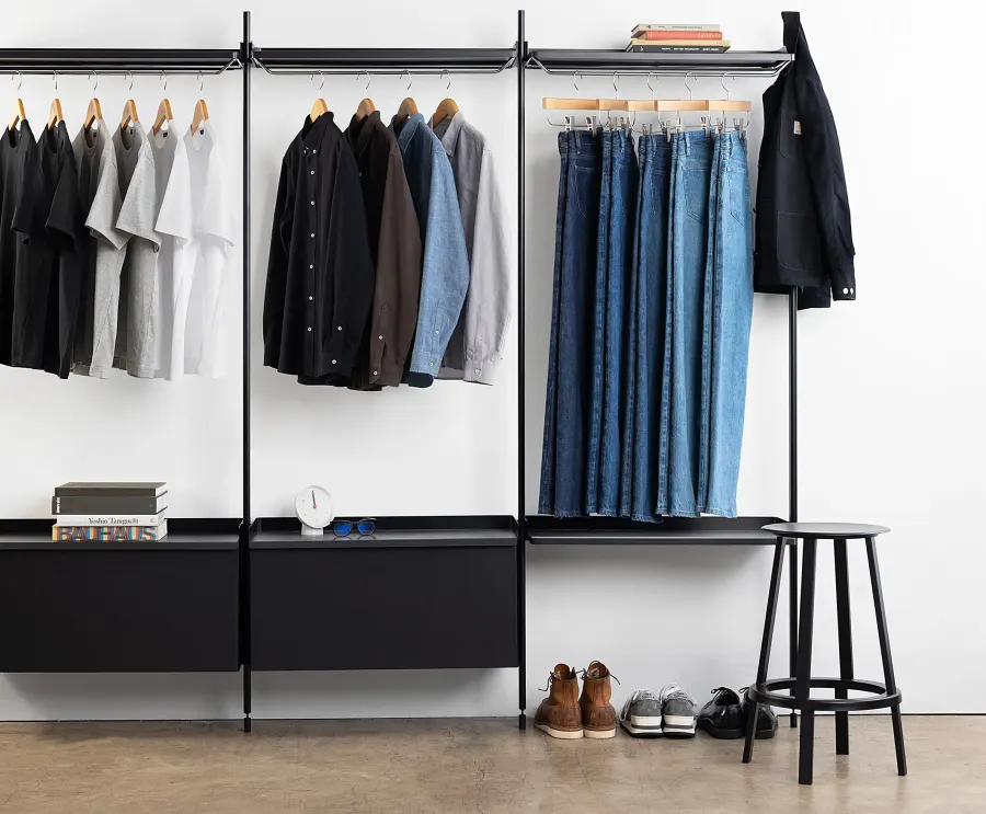 T-shirts, button-up shirts, and blue jeans hang on black clothing racks against a white wall.