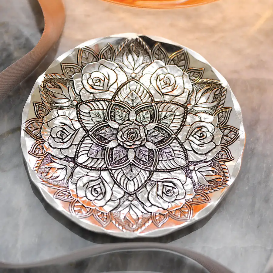 A round hand-hammered aluminum coaster embossed with a symmetrical mandala of rose blossoms and leaves, photographed at an angle on a marble surface.