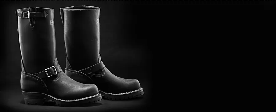 A pair of tall black leather engineer boots with buckles and lug soles, dramatically lit against a near-black background.