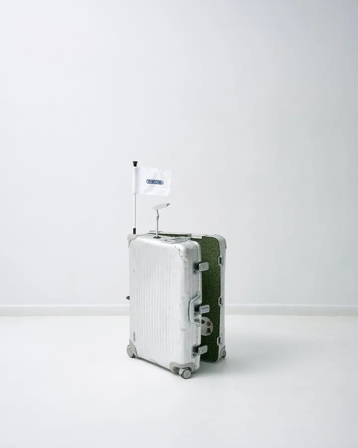 A silver aluminum rolling suitcase with one side finished in artificial putting-green turf, fitted with a small flag and putter, standing against a plain white studio background.