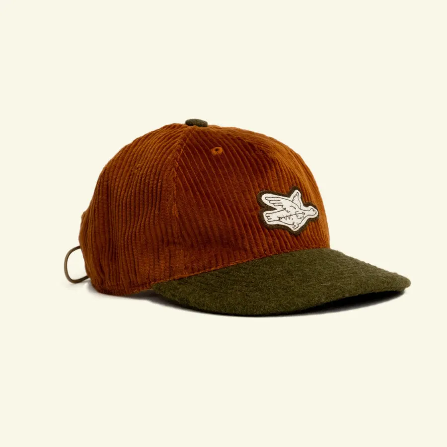 A rust-orange corduroy baseball cap with an olive wool brim and a white bird patch on the crown, photographed against a cream background.