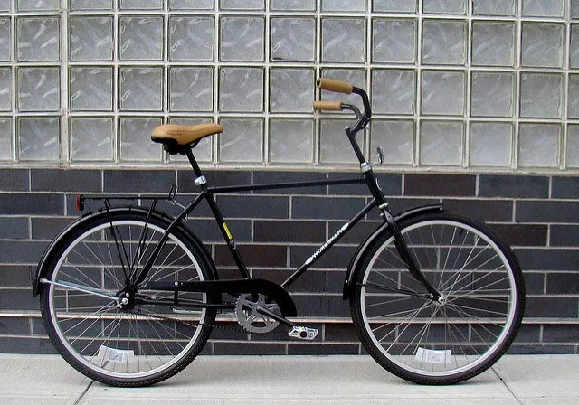 A black Worksman cruiser-style bicycle with a curved BMX-style handlebar parked against a glass-block and brick wall.